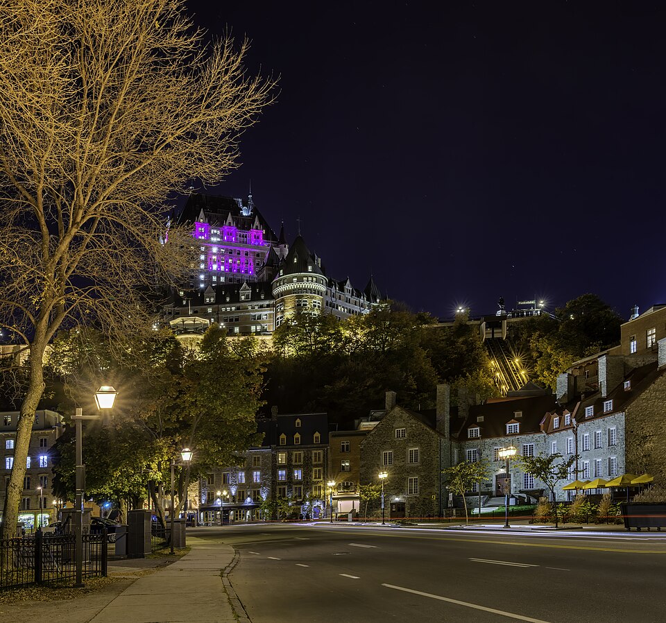 Une vue du Vieux-Québec dans la ville de Québec la nuit - les bâtiments gravissent une haute colline, surmontée par le Fairmont éclairé par une lumière magenta.